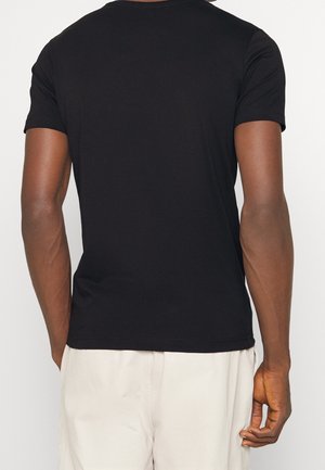 Mann, der ein schlichtes schwarzes kurzärmeliges T-Shirt und hellbeige Hosen trägt, von hinten vor einem schlichten Hintergrund gezeigt.