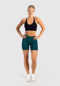 Zwarte sportbeha met gathered detail, gecombineerd met tealkleurige high-waisted short. Model staat met handen op de heupen, met witte sneakers aan.