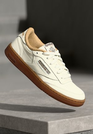 Biały tenis Reebok Classic z brązową podeszwą i beżową wyściółką, umieszczony na kamiennej powierzchni na tle ciemnego rozmytego tła.