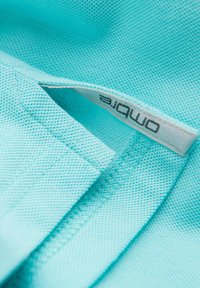 Světle tyrkysová látka s texturovaným povrchem; obsahuje bílou etiketu s nápisem "Ombre" vytištěným šedě. Zaměřte se na tkaní a švy.