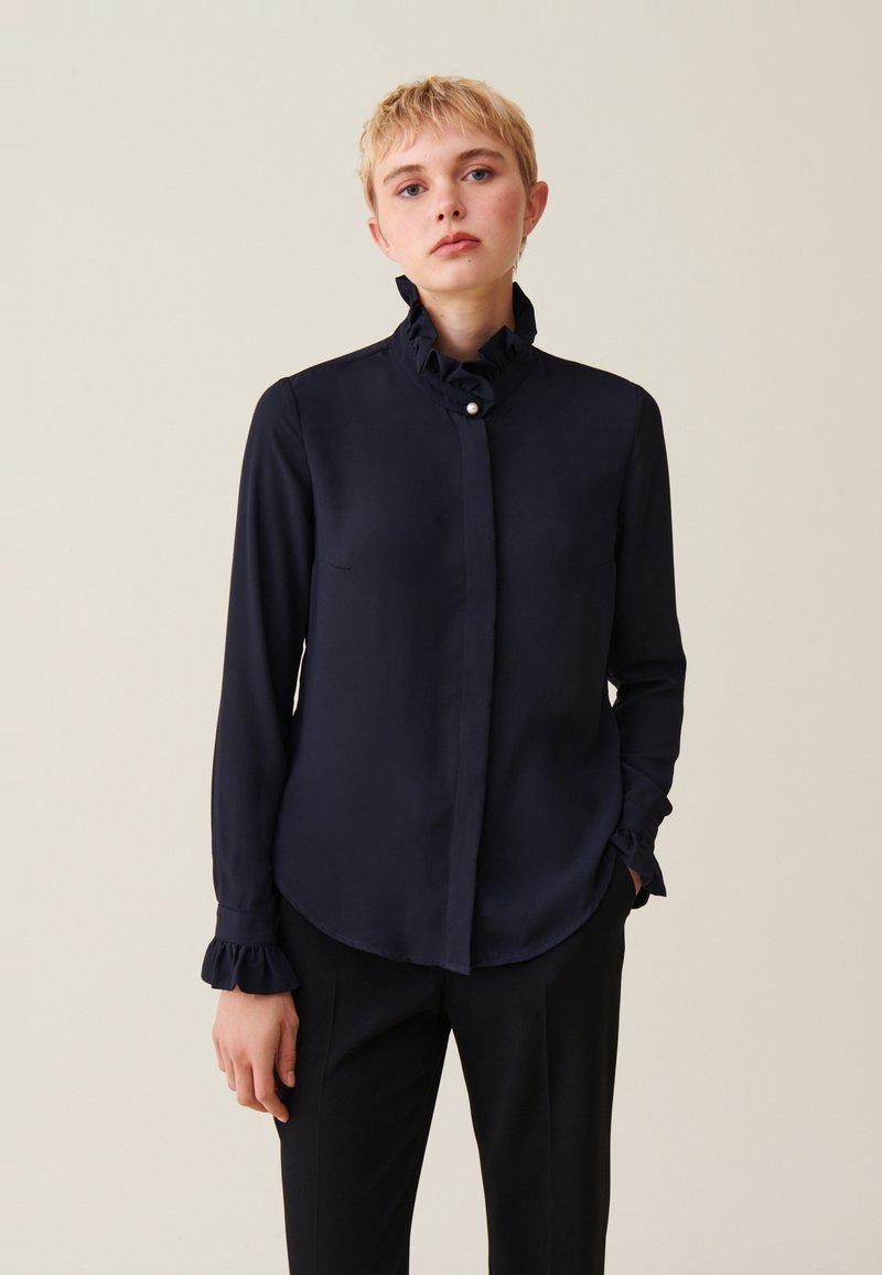 Blusa a maniche lunghe blu navy con colletto arricciato, chiusura a bottoni e polsini arricciati. Tessuto liscio con una vestibilità su misura. Nessun motivo visibile.