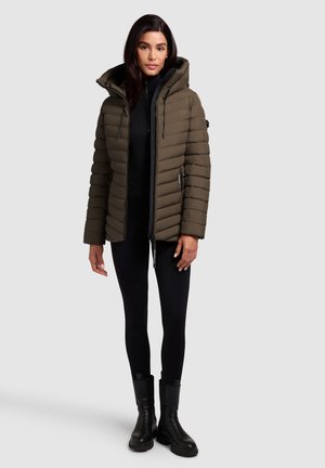 Woman wearing an olive green quilted puffer jacket, black leggings, and black ankle boots, standing against a plain light gray background.