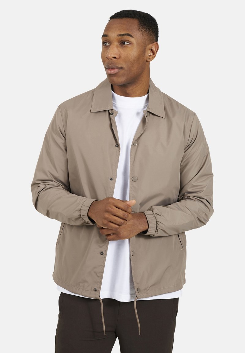 Jasnobeżowa kurtka bomberka z kołnierzem, z zatrzaskami, elastycznymi mankietami oraz ściągaczem w pasie. Nosić ją można na białą koszulkę.