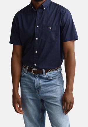 Un homme porte une chemise bleu marine à manches courtes avec une poche poitrine et un jean bleu clair, accessoirisé d'une ceinture sombre à motifs.