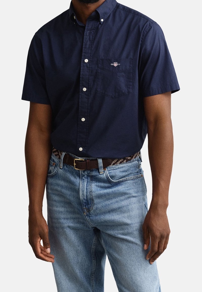 Un homme porte une chemise bleu marine à manches courtes avec une poche poitrine et un jean bleu clair, accessoirisé d'une ceinture sombre à motifs.