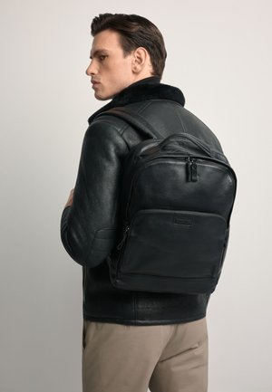 Czarny skórzany plecak o gładkiej fakturze, zaokrąglonym kształcie i kieszeniach na zamek. Noszony na czarnej skórzanej kurtce.