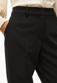 Kvinne i høytaljede svarte skræddersydde bukser med beltehemper, sidelommer, og en kremfarget bluse med rynkede mansjetter, hånd i lommen.