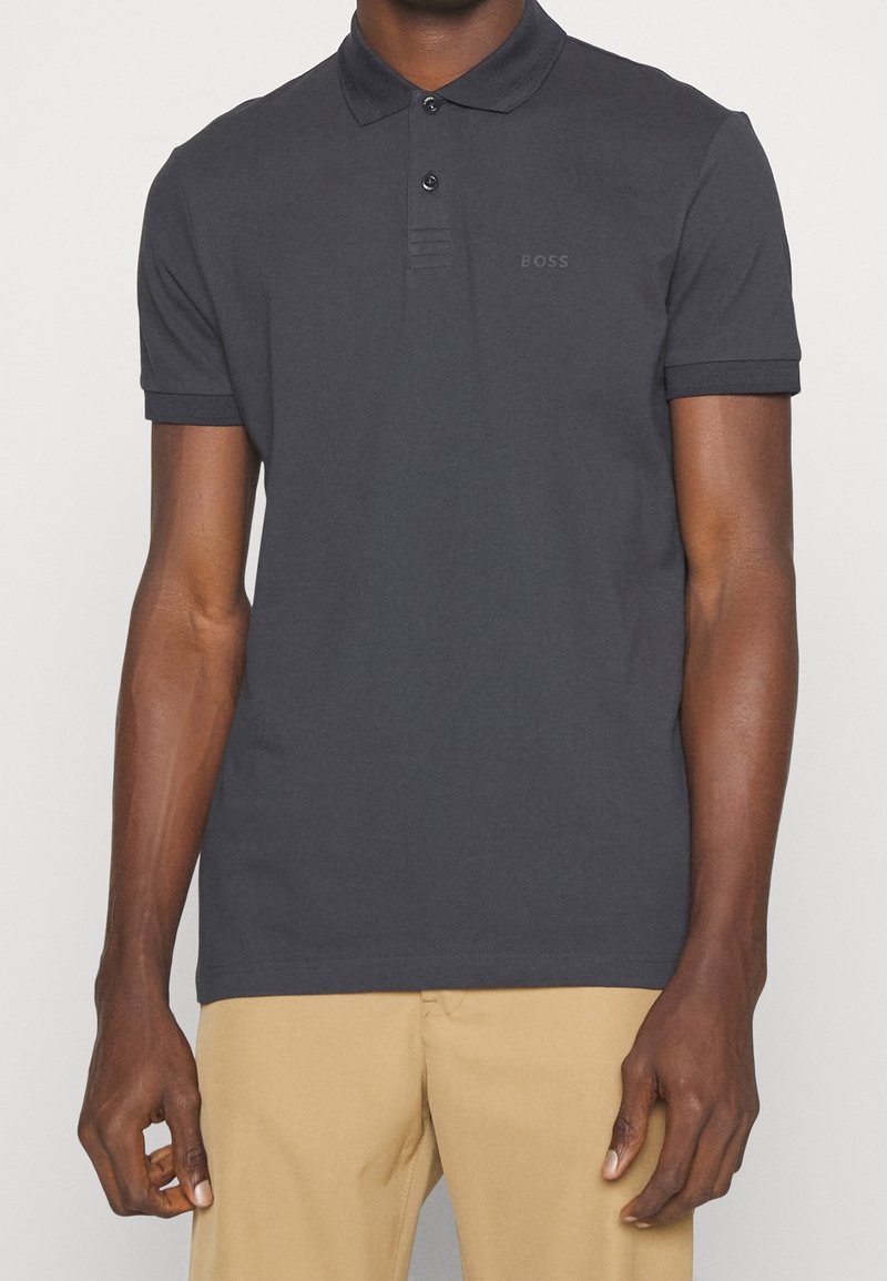Homme portant un polo gris foncé à manches courtes avec le logo "BOSS" sur la poitrine et un pantalon beige, debout devant un fond clair uni.