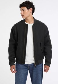 Giacca bomber grigio scuro realizzata in un tessuto morbido. Presenta una zip frontale, polsini a coste e una tasca laterale con zip. Abbinata a una t-shirt bianca.