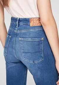 Blå denimjeans med hög midja, med en brun läderlapp märkt "PEPE JEANS." Inkluderar två bakfickor och sömdekor. 