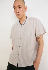 Chemise à manches courtes, col classique en beige avec de fines rayures verticales, dotée de boutons sur le devant et d'une poche poitrine unique. Texture douce.