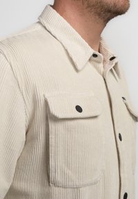 Beige Cordjacke mit Kragen, zwei aufgesetzten Taschen mit schwarzen Knöpfen und einem strukturierten Rippmuster.