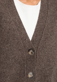 Hnědý pletený kardigan s hlubokým výstřihem do V, se dvěma černými knoflíky a strukturovanou látkou s fleky světlejších barev. Nositý přes bílý top.