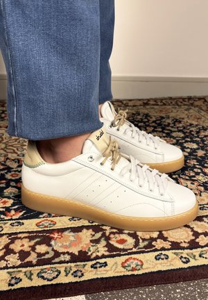 Persoon die witte sneakers draagt met beige accenten en gumzolen, staand op een patroonmatig tapijt waarbij blauwe jeans zichtbaar zijn.