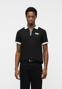 Νεαρός άνδρας φοράει μαύρο πόλο με λευκό γιακά και λευκές λεπτομέρειες στα μανίκια, στέκεται μπροστά σε απλό φωτεινό φόντο.