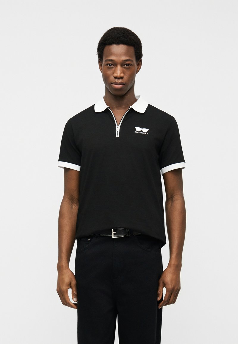 Νεαρός άνδρας φοράει μαύρο πόλο με λευκό γιακά και λευκές λεπτομέρειες στα μανίκια, στέκεται μπροστά σε απλό φωτεινό φόντο.