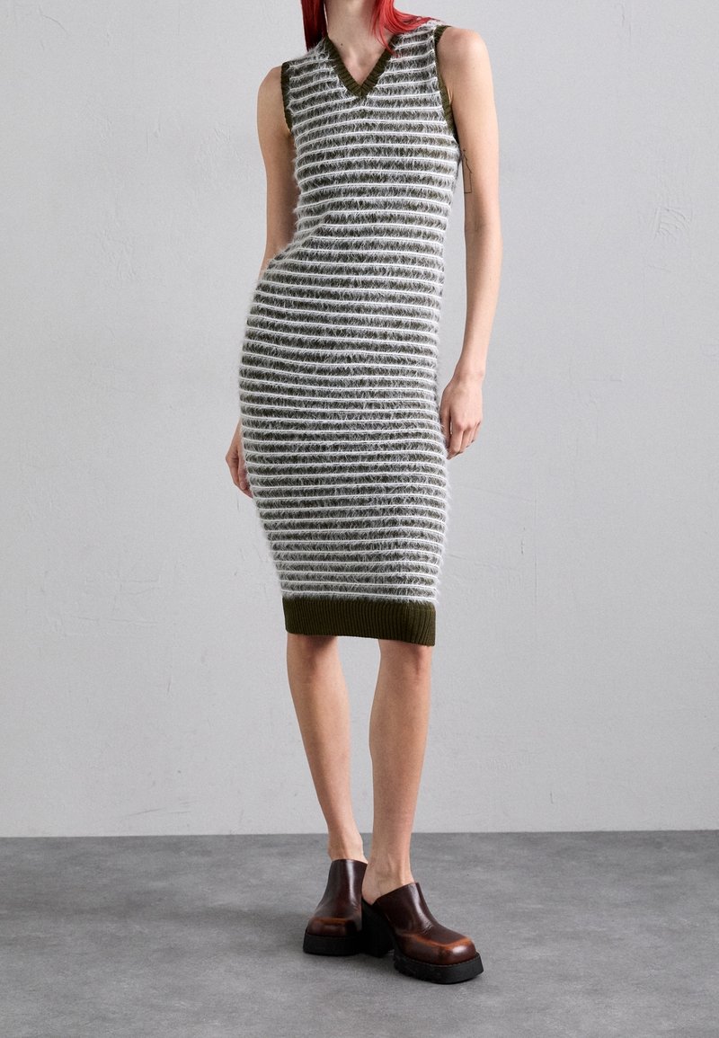 Personne portant une robe en maille duveteuse sans manches, longueur genou, à rayures horizontales et des chaussures compensées marron, debout sur un sol gris.