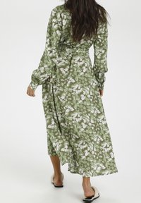 Kobieta idąca w stronę wyjścia w długiej zielono-białej sukience w kwiaty i beżowych sandałach wsuwanych na jednolitym białym tle.