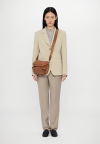 Beige přizpůsobený blazer přes šedou košili s knoflíky, spárovaný se světle béžovými kalhotami a černými mokasíny; hnědá kabelka přes rameno s řetízkovým detailem.