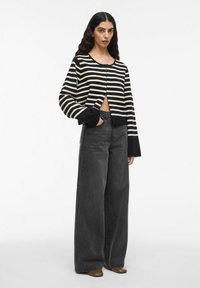 Donna con lunghi capelli scuri, vestita con un maglione corto a righe bianche e nere e jeans grigi scuri a vita alta e gambali larghi, abbinati a scarpe marroni.