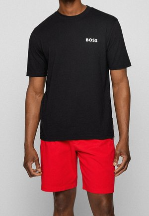 Mand iført en sort kortærmet T-shirt med teksten "BOSS" på brystet og knaldrøde knælange shorts mod en ensfarvet baggrund.