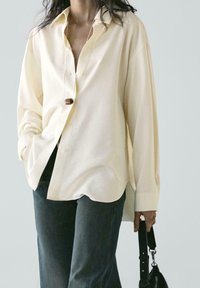 Camicia oversize color crema con chiusura frontale a bottoni, maniche lunghe e orlo leggermente arrotondato. Abbinata a denim scuro e una borsa nera.