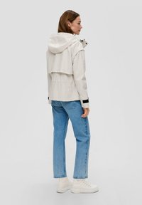 Hellbeige Regenjacke mit Kapuze, elastischem Taillenbund und schwarzen Akzenten. Trägt man mit blauen Jeans und weißen Sneakers. Rückansicht gezeigt.