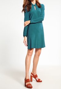 Teal klänning med långa ärmar, nyckelhålskärning och samlingsdetaljer vid ärmsluten. Matcher med bruna mocka blockklack-sandaler.