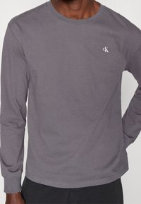 Pitkähihaisen harmaan puuvillapaidan, jossa on pyöreä kaula-aukko, etupuolella pieni valkoinen "CK"-logo vasemmalla rintapielessä. Sujuva rakenne ja istuva muotoilu.