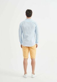 Chemise à manches longues bleu clair associée à un short jaune ; des baskets blanches complètent le look. La chemise a un ourlet arrondi, les shorts sont à mi-cuisse.