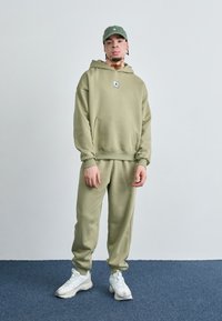 Olivengrøn hoodie og sweatpants sæt, blødt stof, kængurulomme, logo lap på brystet, parret med hvide sportsko.