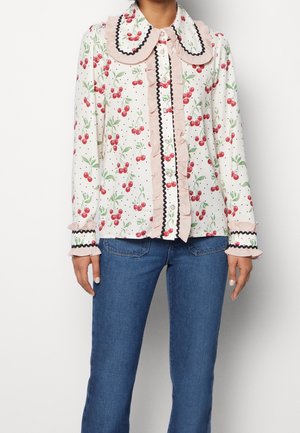 Blouse blanche à manches longues avec imprimé cerises rouges, col et poignets à volants roses, bordure noire, portée avec un jean bleu.
