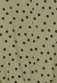 Tessuto verde oliva con pois neri, con un dettaglio logo visibile. La trama appare liscia e leggera.