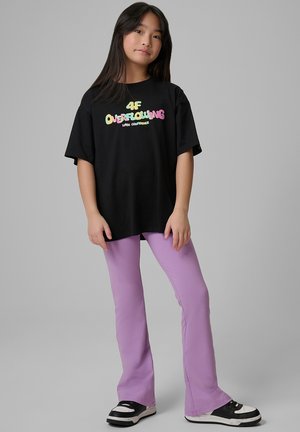 Młoda dziewczyna o długich ciemnych włosach, ubrana w czarny, oversize’owy t-shirt z kolorowym napisem i lawendowe rozszerzane spodnie, stojąca na jednolitym szarym tle.