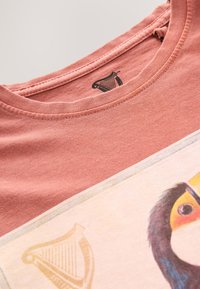 Close-up of a pink T-shirt neckline with a printed label and partial graphic of a colorful bird and gold harp symbol.