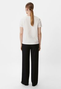 Weißes Baumwoll-T-Shirt mit kurzen Ärmeln und lockerer Passform, kombiniert mit schwarzen weit geschnittenen Hosen. Hintergrund ist schlicht weiß.