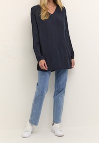 Frau trägt eine lockere, langärmlige marineblaue Tunika, hellblaue gerade Jeans, weiße Sneaker und steht vor einem schlichten hellen Hintergrund.