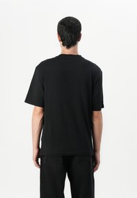 Black cotton t-shirt with a relaxed fit, short sleeves, and a crew neckline, displayed from the back, paired with black denim pants.