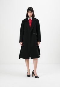 Donna che indossa un lungo cappotto nero, un maglione rosso con colletto bianco e tacchi neri, in piedi contro uno sfondo bianco semplice.