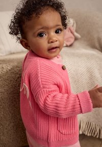 Cardigan rosa lavorato a maglia con texture a coste, con bottone rotondo in legno e tasche. Indossato sopra una maglietta rosa chiaro.