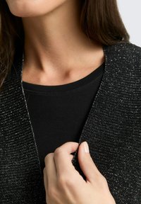 Black textured cardigan with metallic thread accents, worn over a fitted black top. Close-up of hand holding the cardigan's edge.