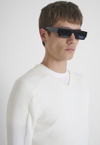 Occhiali da sole neri a forma rettangolare con una montatura spessa, indossati da una modella in un maglione bianco a maglia, con una texture a coste e design a scollo a V.