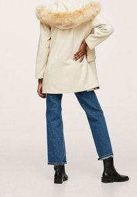 Abrigo beige con capucha de pelo, corte recto y dos bolsillos. Combinado con unos vaqueros azules y botas negras de tobillo, mostrando un atuendo casual.