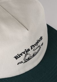 Béžová kšiltovka s tmavě zeleným kšiltem, s vyšitým nápisem "Rivvia Projects" a ilustrací hada na přední straně. Texturovaná tkanina.