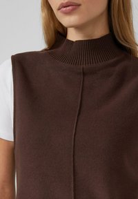 Brauner ärmelloser Pullover mit hohem, geripptem Kragen, weicher Textur und zentraler Nahtdetail. Trägt über einem weißen Kurzarmshirt.