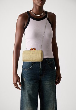 Donna che indossa un top bianco con rifiniture nere, jeans di denim a gamba larga, collana di conchiglie e una piccola borsa a tracolla beige intrecciata con chiusura in ambra.