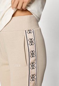 Μπεζ φόρμες με ριχτό παντελόνι, που διαθέτουν μαύρες λεπτομέρειες στο πλαϊνό πάνελ και ένα μικρό λογότυπο "GUESS" μπροστά.