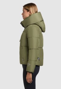 Olijfgroene pufferjack met een hoge kraag en een afneembare capuchon. Voorzien van een gestikt patroon, een ritssluiting aan de voorkant en twee zijzakken.