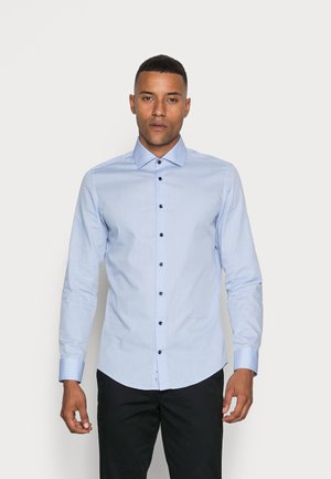 Mężczyzna ubrany w jasnoniebieską koszulę na guziki i czarne spodnie, stojący na jednolitym jasnym tle.