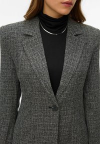 Szary, teksturowany żakiet z wyraźnymi ramionami, zapinany na jeden czarny guzik, noszony na czarnym golfie. Akcent w postaci metalicznego srebrnego naszyjnika.